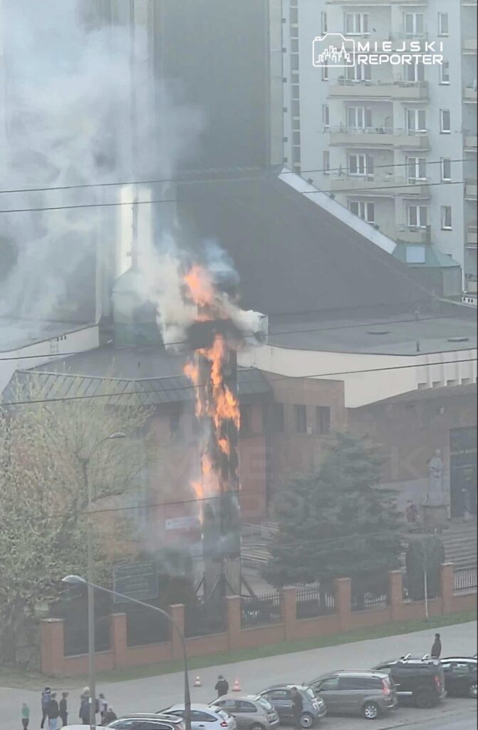 Płomienie wydobywające się z dachu budynku, w otoczeniu parku i zaparkowanych samochodów.