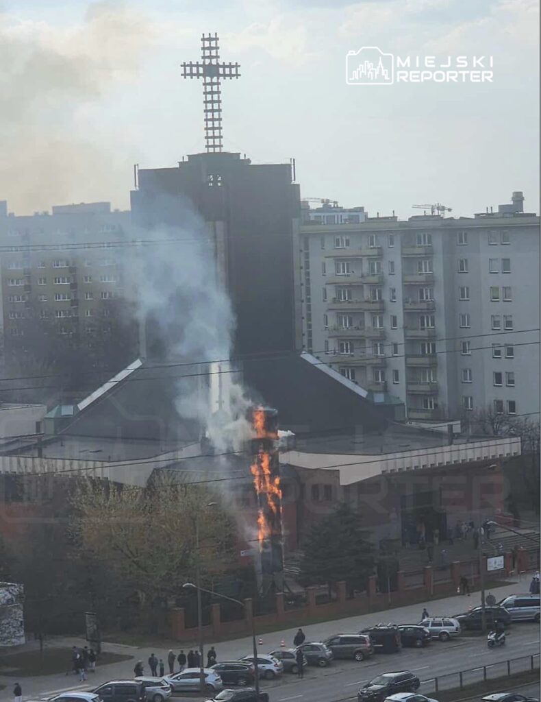 Kościół z dymem i płomieniami wydobywającymi się z komina, w tle bloki mieszkalne i zgromadzeni ludzie.
