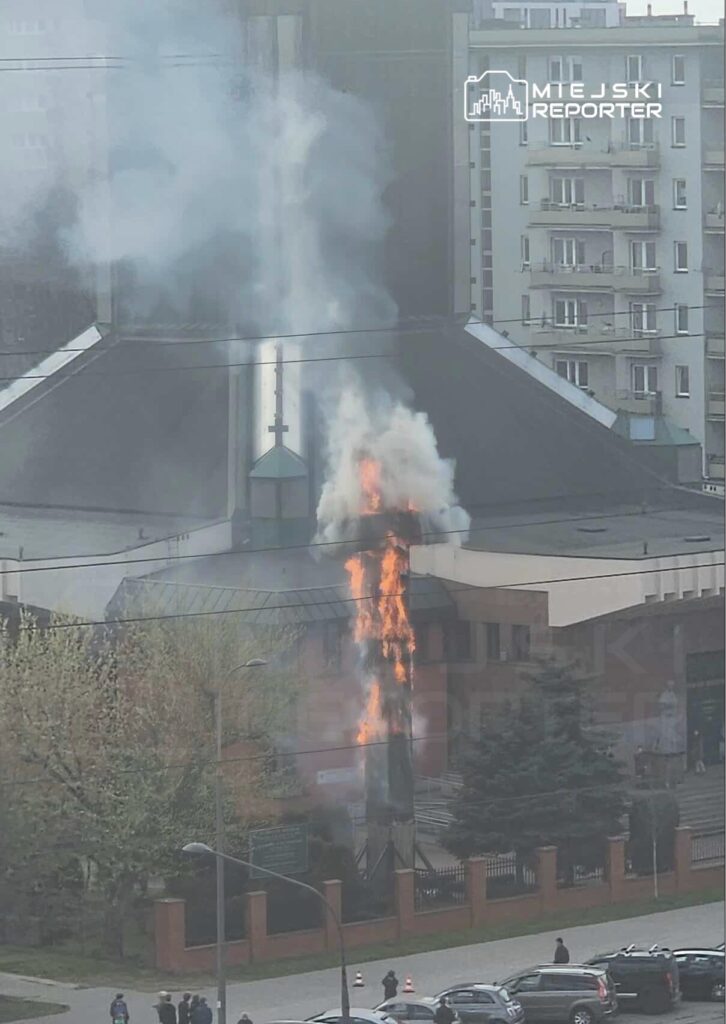Płomienie wydobywające się z komina budynku, otoczone dymem, w tle widoczne samochody i drzewa.