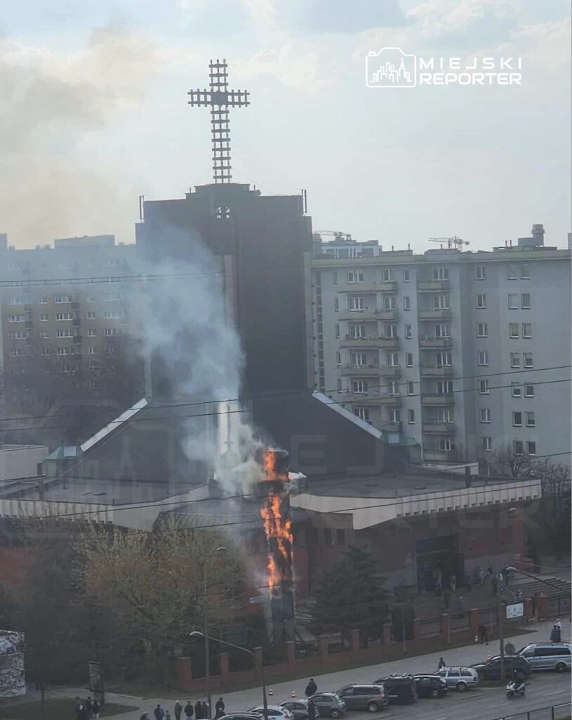 Pożar na dachu budynku sakralnego, z którego wydobywa się dym i płomienie, w pobliżu mieszkańców obserwujących sytuację.