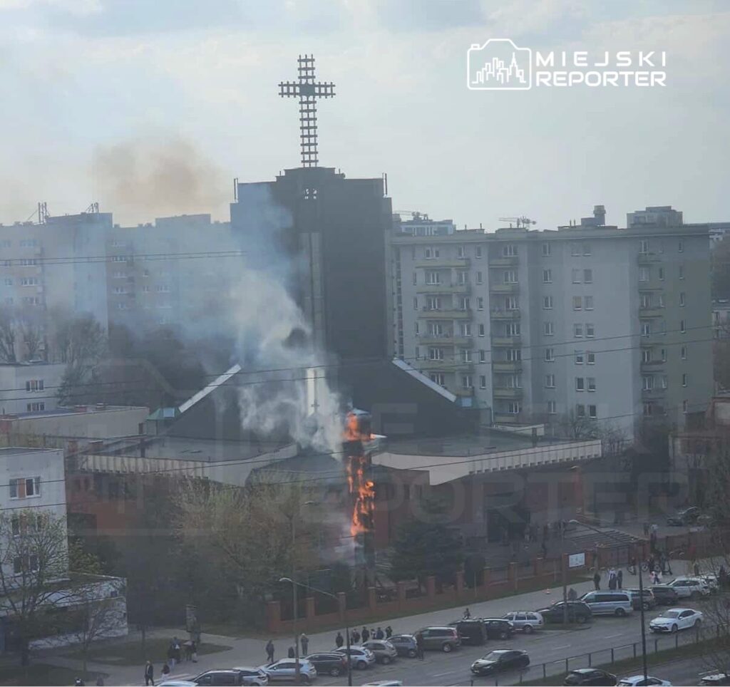 Kościół z dymem i płomieniami wydobywającymi się z dachu, w tle bloki mieszkalne i przechodnie na ulicy.