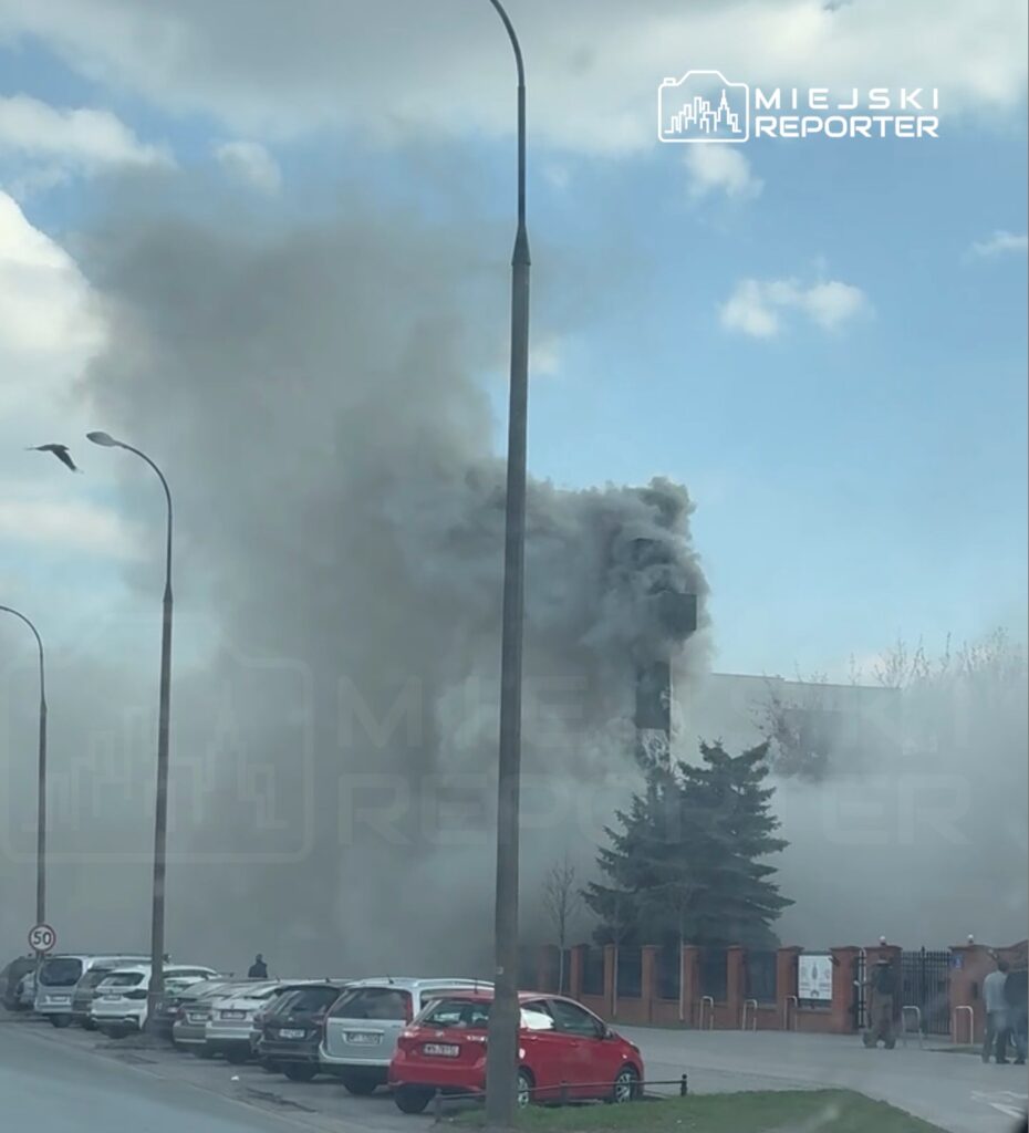 Gęsty dym unoszący się z budynku w miejskim otoczeniu, w pobliżu zaparkowanych samochodów.