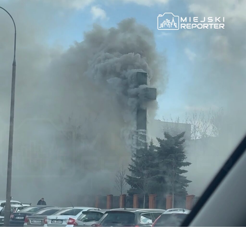 Gęsty dym unoszący się z wysokiego krzyża w otoczeniu samochodów na parkingu.