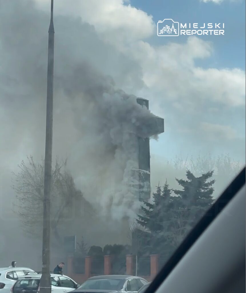 Gęsty dym wydobywający się z budynku w otoczeniu drzew i zaparkowanych samochodów.