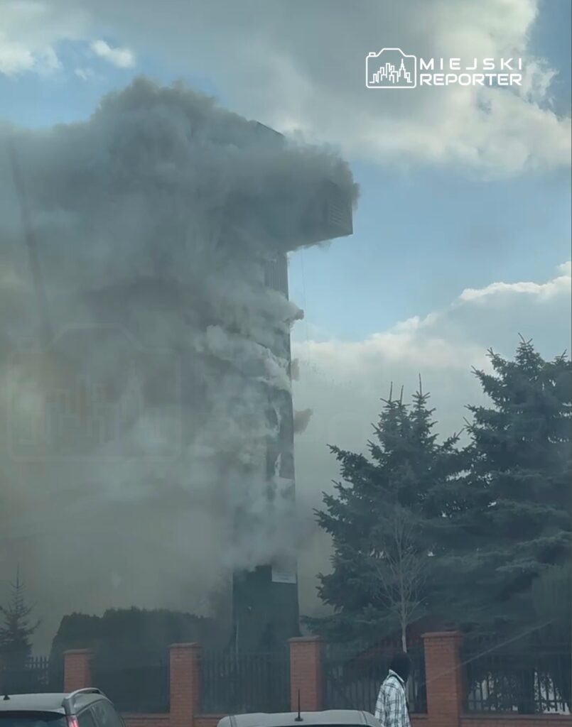 Wysoka wieżowca wydobywa się gęsty dym, a w tle widać drzewa oraz zaparkowane samochody.