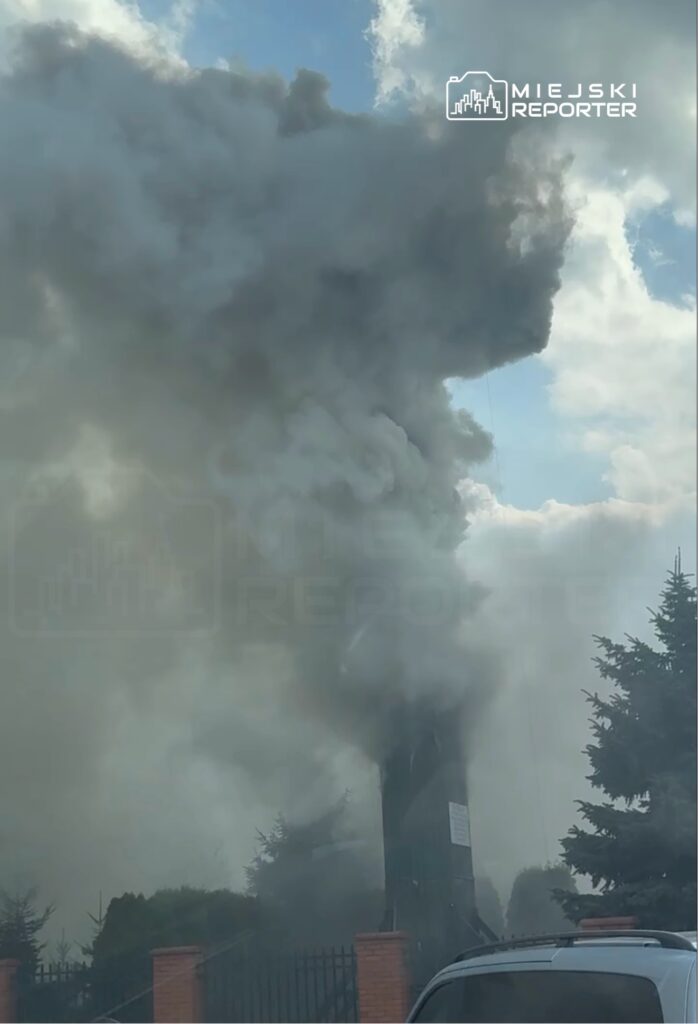 Gęsty, szary dym unoszący się z budynku, otoczonego zielonymi drzewami i ogrodzeniem.