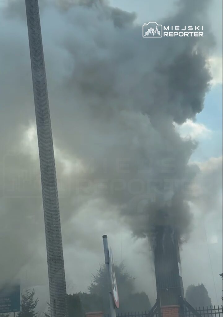 Gęsty dym wydobywający się z wysokiego komina w miejskim otoczeniu, w tle widoczne drzewa i znak drogowy.