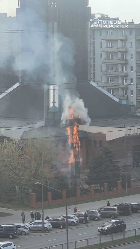 W budynku kościoła unosi się dym, a z jego dachu wydobywają się płomienie. Grupa osób obserwuje zdarzenie z chodnika.