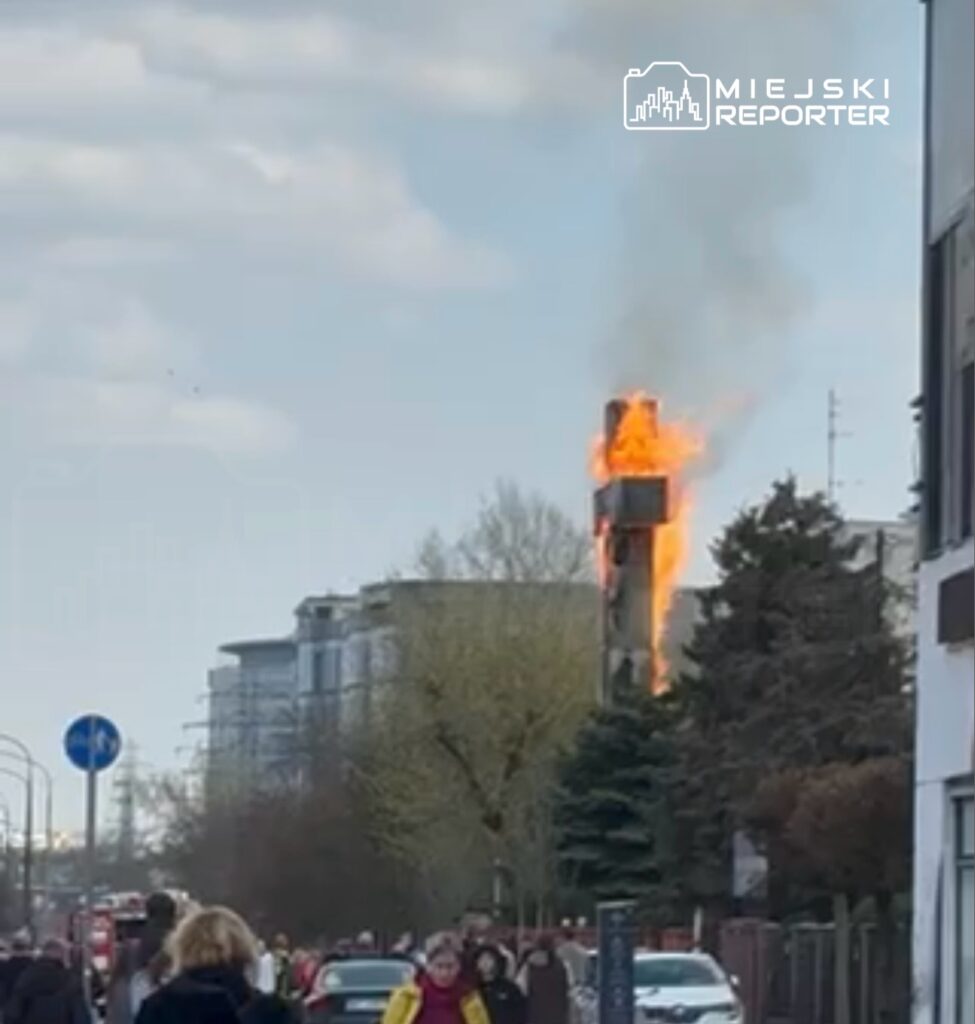 Wysoka wieża w płomieniach, otoczona ludźmi obserwującymi zdarzenie na ulicy.