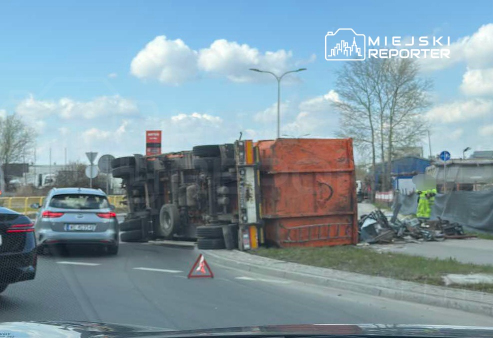 Przewrócony ciężarówka na drodze, obok stojące samochody i znak ostrzegawczy. W tle widoczna infrastruktura miejska.