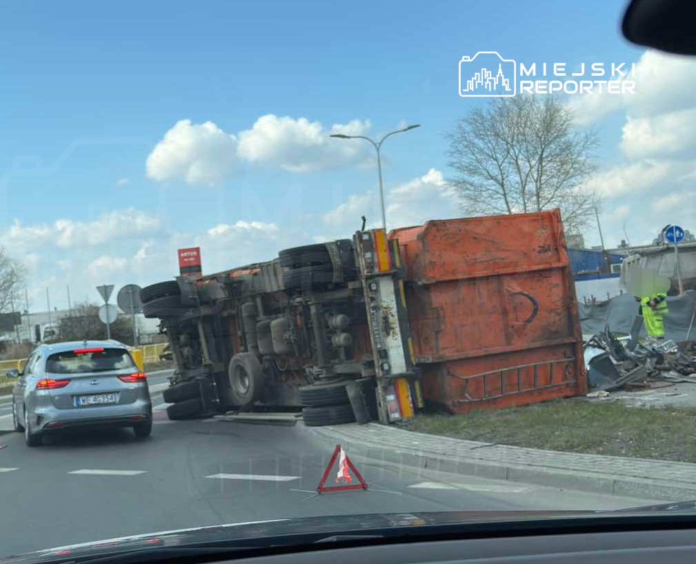Czerwony ciężarówka leży na boku na rondzie, obok stoi samochód osobowy, a w tle widoczny pracownik w kamizelce odblaskowej.