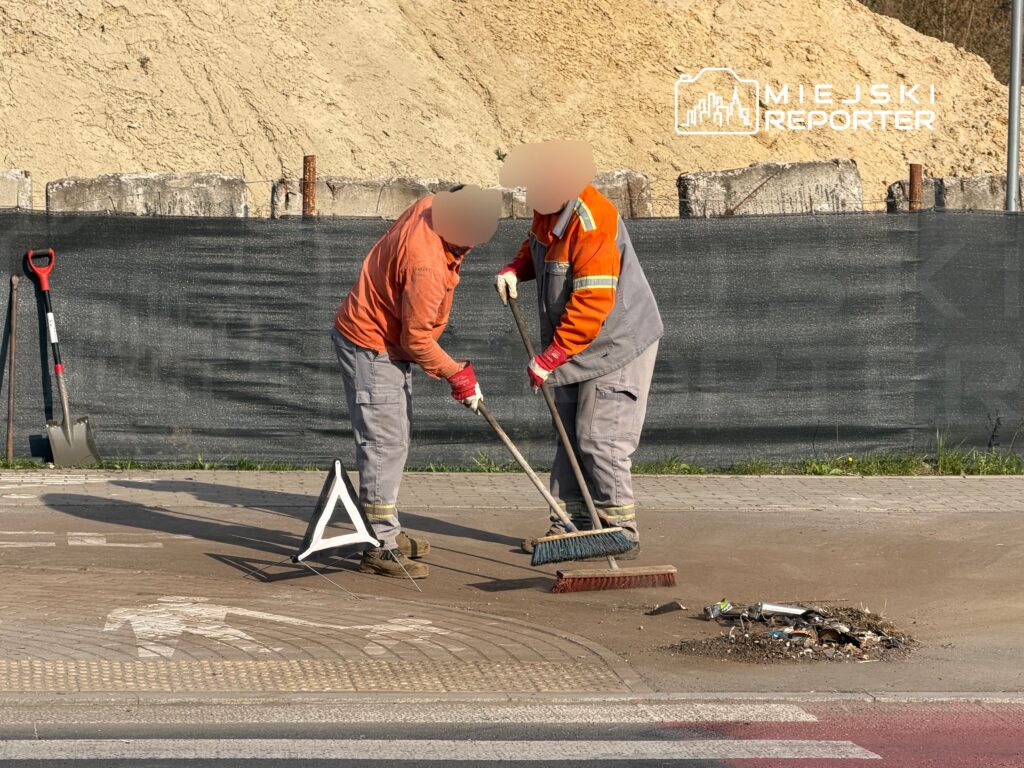 Dwaj pracownicy w pomarańczowych kurtkach zamiatają chodnik obok sterty odpadków w terenie budowlanym.