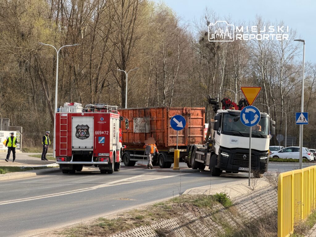 Strażacy i policjanci monitorują sytuację przy drodze, gdzie ciężarówka z kontenerem jest holowana przez inny pojazd.