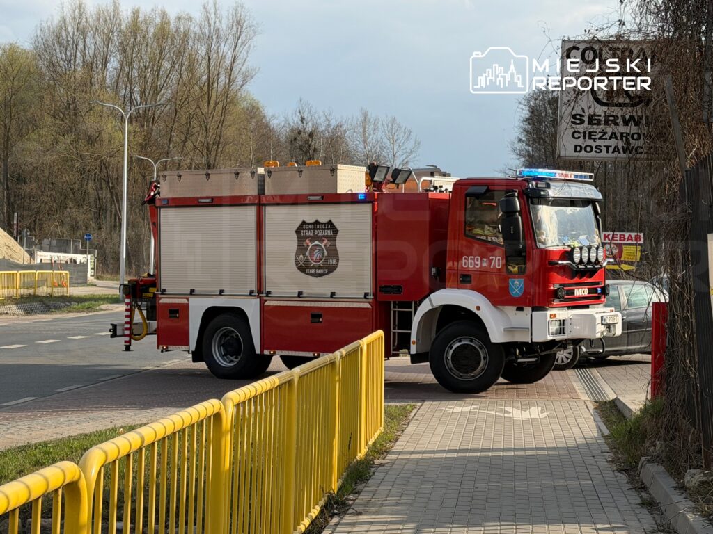 Czerwony wóz strażacki z włączonymi sygnalizatorami zatrzymany na skrzyżowaniu obok żółtej bariery ochronnej.