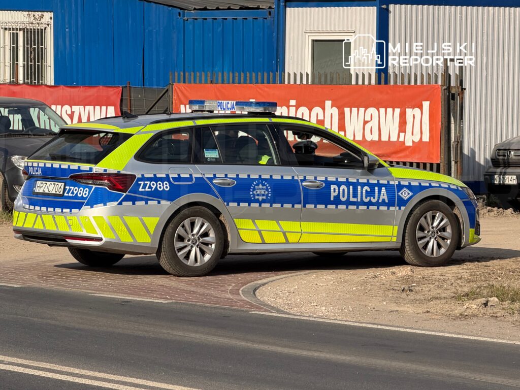 Radiowóz policyjny z oznaczeniami i numerem ZZ988 zaparkowany na poboczu drogi, w tle widoczny niebieski budynek i reklama.