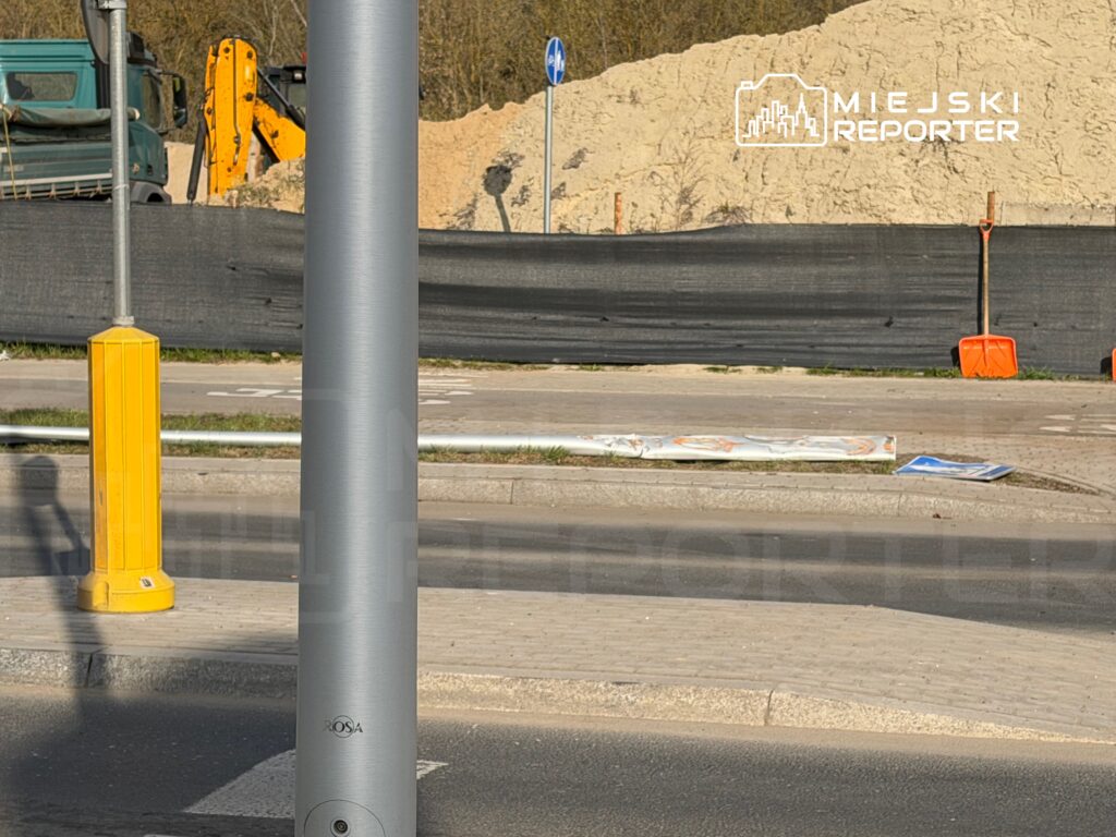 Srebrny słup latarni ulicznej stoi obok żółtego słupka, w tle widoczna budowa z koparką i ogrodzeniem.