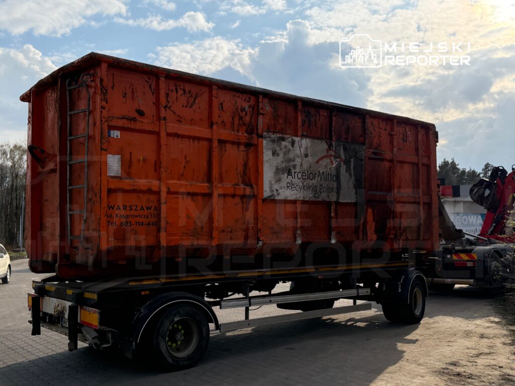 Pomarańczowy kontener na odpady umieszczony na ciężarówce, w tle chmury i drzewa. Na kontenerze widoczny napis „ArcelorMittal Recykling Polska”.