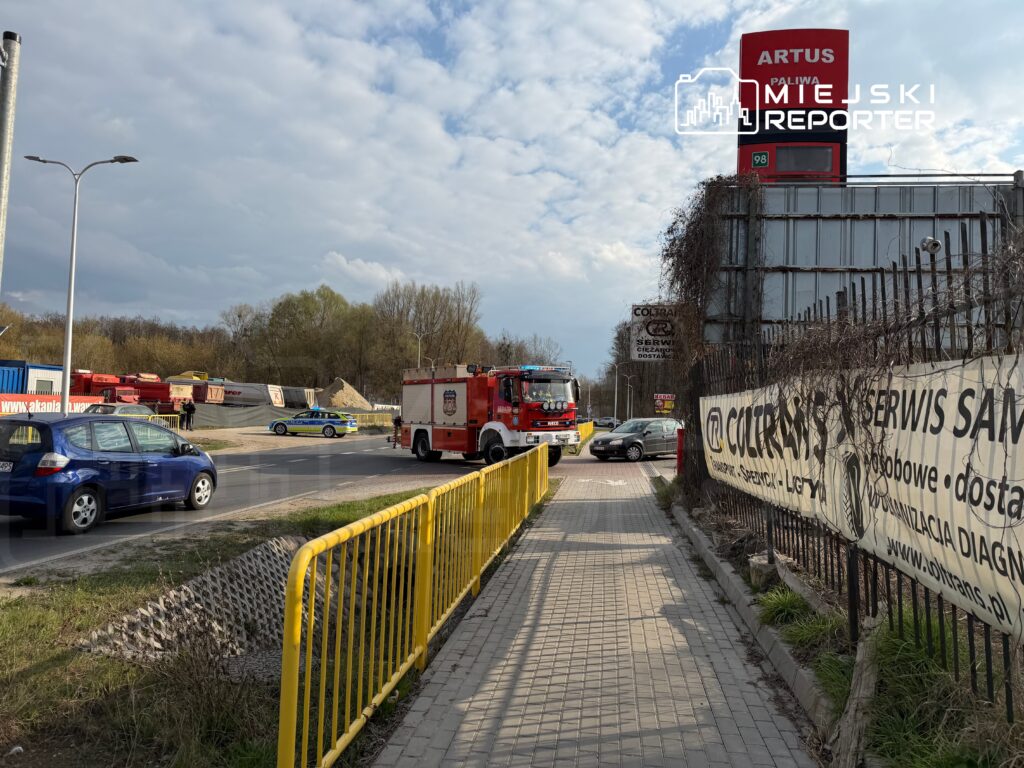 Straż pożarna z włączonymi sygnałami świetlnymi zatrzymana przy ruchliwej ulicy obok żółtej bariery ochronnej.