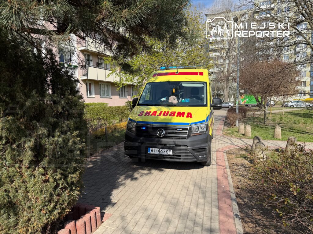 Karetka pogotowia zaparkowana na chodniku w pobliżu budynków mieszkalnych, otoczona drzewami i krzewami.