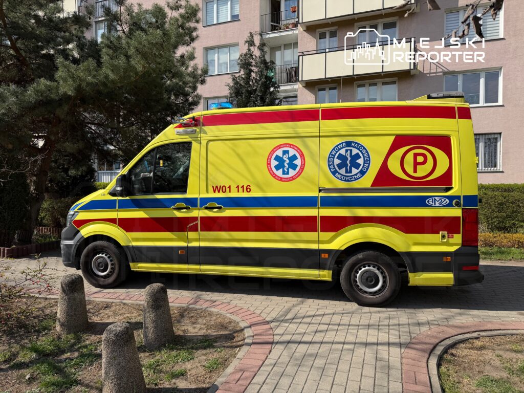 Karetka pogotowia ratunkowego zaparkowana na chodniku przed budynkiem mieszkalnym, otoczona zielenią.