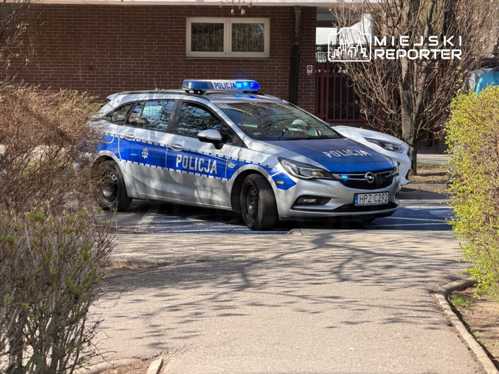 Policyjny samochód osobowy z niebieskimi światłami zaparkowany na chodniku w otoczeniu drzew i krzewów.