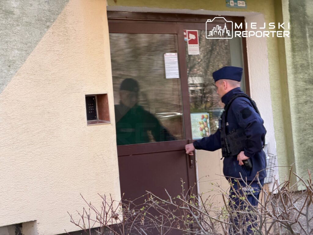 Policjant w mundurze otwiera drzwi do budynku, trzymając w ręku klamkę. W tle widoczna jest zieleń krzewów.