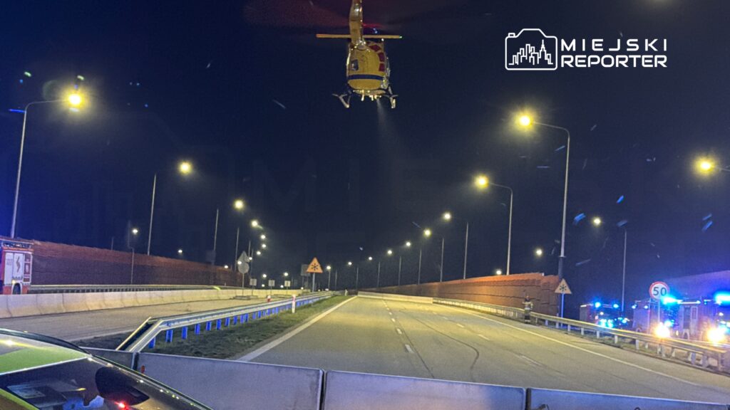 Śmigłowiec ratunkowy unosi się nad oświetloną drogą, na której stoją pojazdy służb ratunkowych.