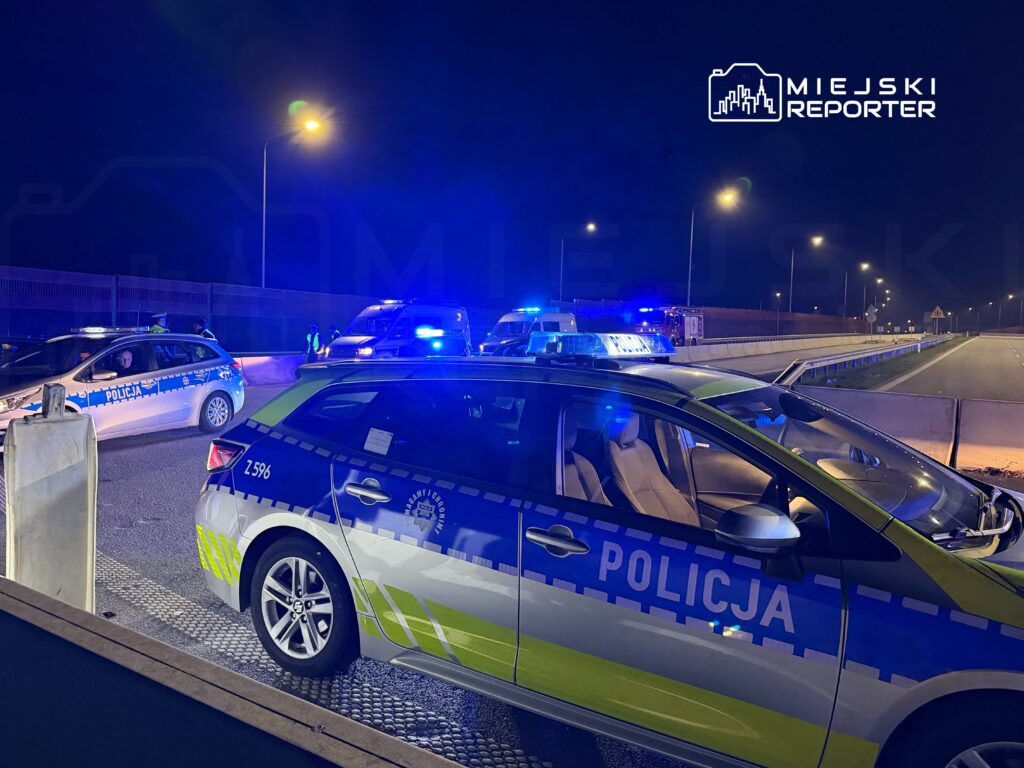 Policyjny radiowóz z włączonymi sygnałami świetlnymi stoi na drodze, w tle widoczny drugi radiowóz oraz pojazdy służb ratunkowych.