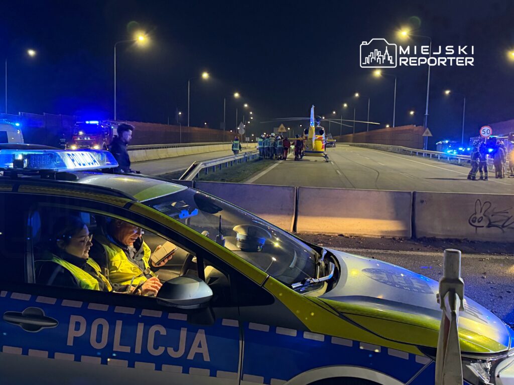 Policjanci w radiowozie analizują sytuację na drodze, gdzie interweniują służby ratunkowe.