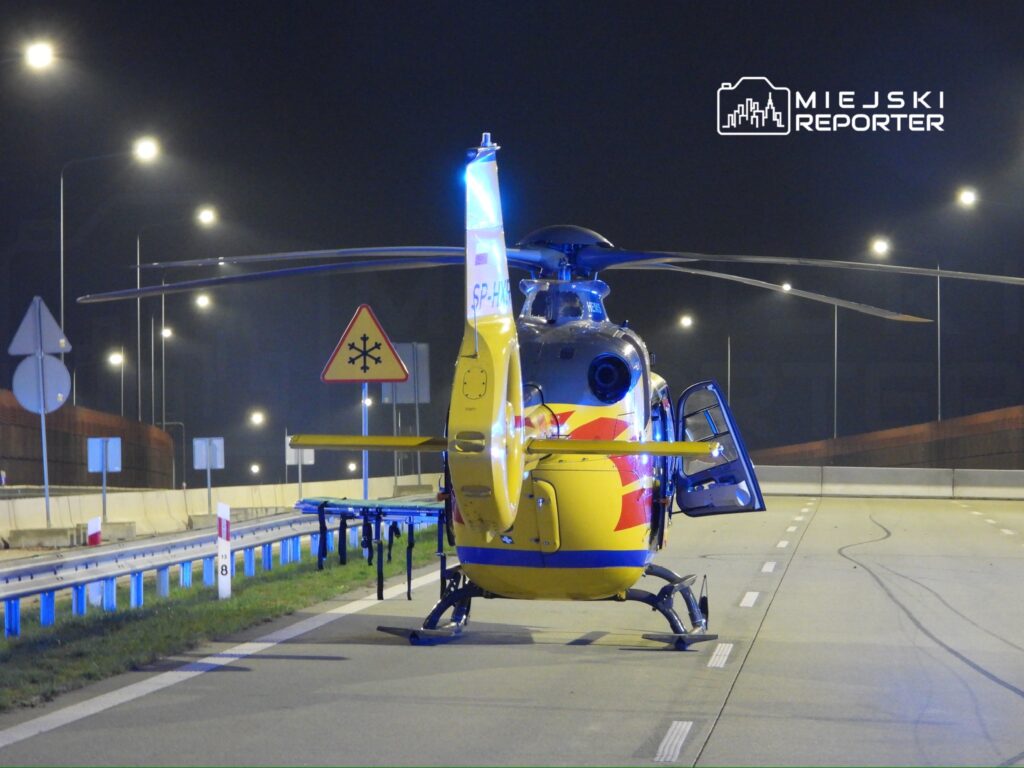 Helikopter ratunkowy z otwartymi drzwiami ląduje na oświetlonej drodze nocą, obok znak ostrzegawczy o śliskiej nawierzchni.