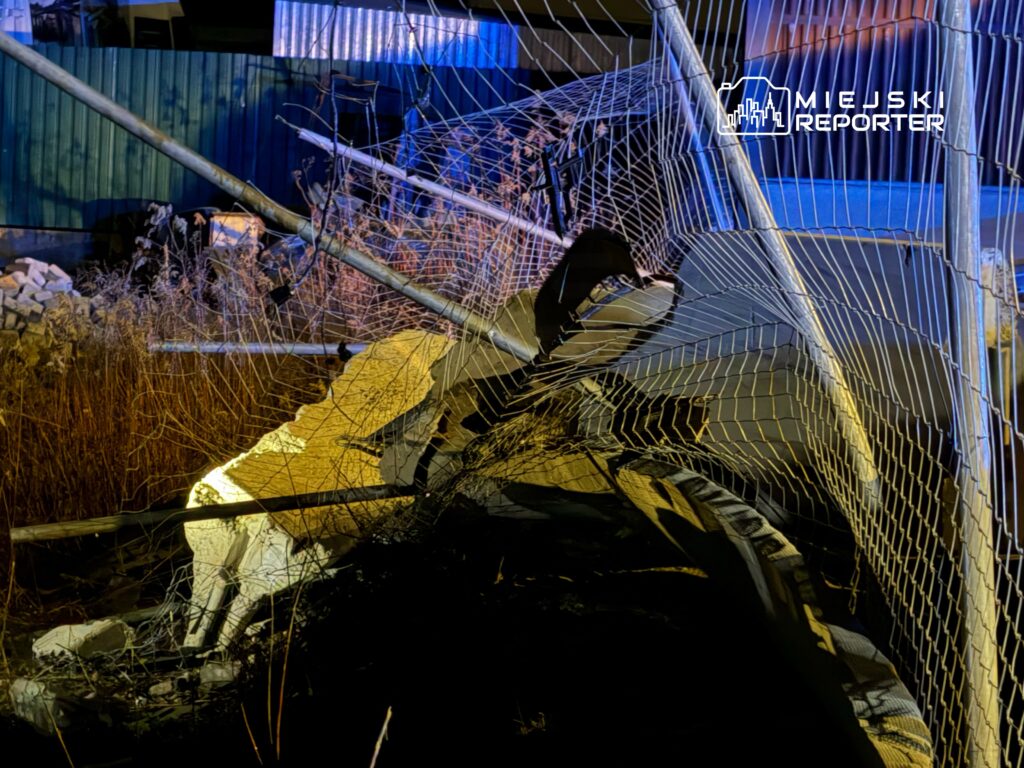 Zniszczona siatka ogrodzeniowa opada na porzucone materiały budowlane w zarośniętym terenie.