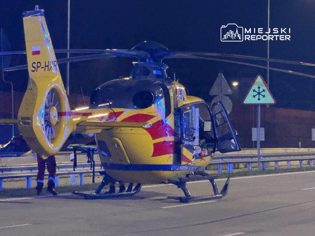 Helikopter ratunkowy z otwartymi drzwiami ląduje na drodze, obok znak drogowy z symbolem śniegu.