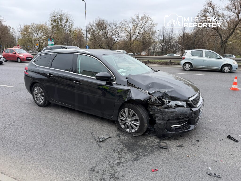 Czarny samochód osobowy z uszkodzoną przednią częścią stoi na jezdni, obok widoczne inne pojazdy i znaki drogowe.