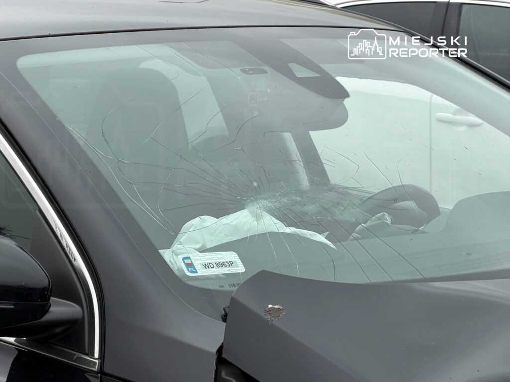Pęknięta szyba czołowa w czarnym samochodzie zaparkowanym na parkingu, w tle widoczny inny pojazd.