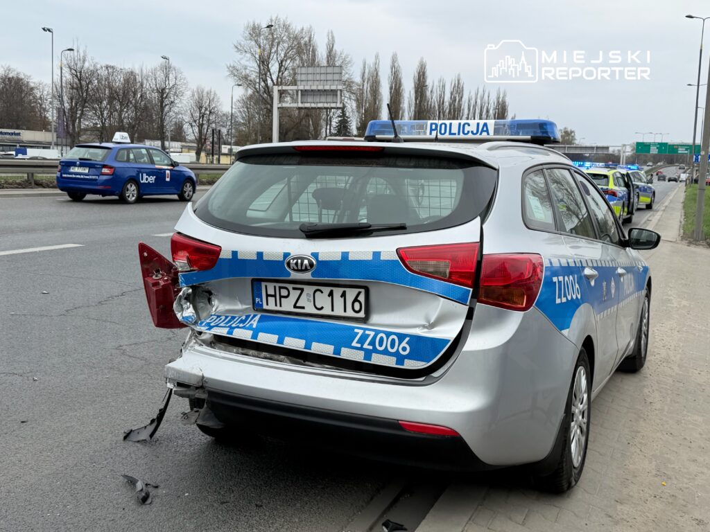 Uszkodzony radiowóz policji z wgniecioną tylną częścią stoi na poboczu drogi, obok przejeżdżający samochód Uber.