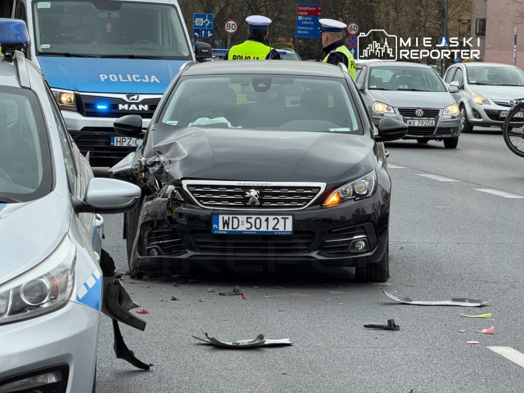 Czarny samochód osobowy z uszkodzoną przednią częścią stoi na drodze, obok policyjny radiowóz z włączonymi sygnałami.