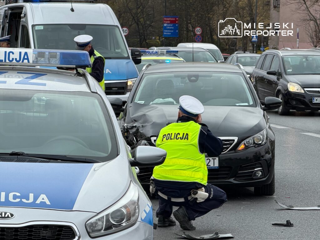 Policjant w kamizelce odblaskowej zbiera informacje przy uszkodzonym czarnym samochodzie na ruchliwej ulicy.