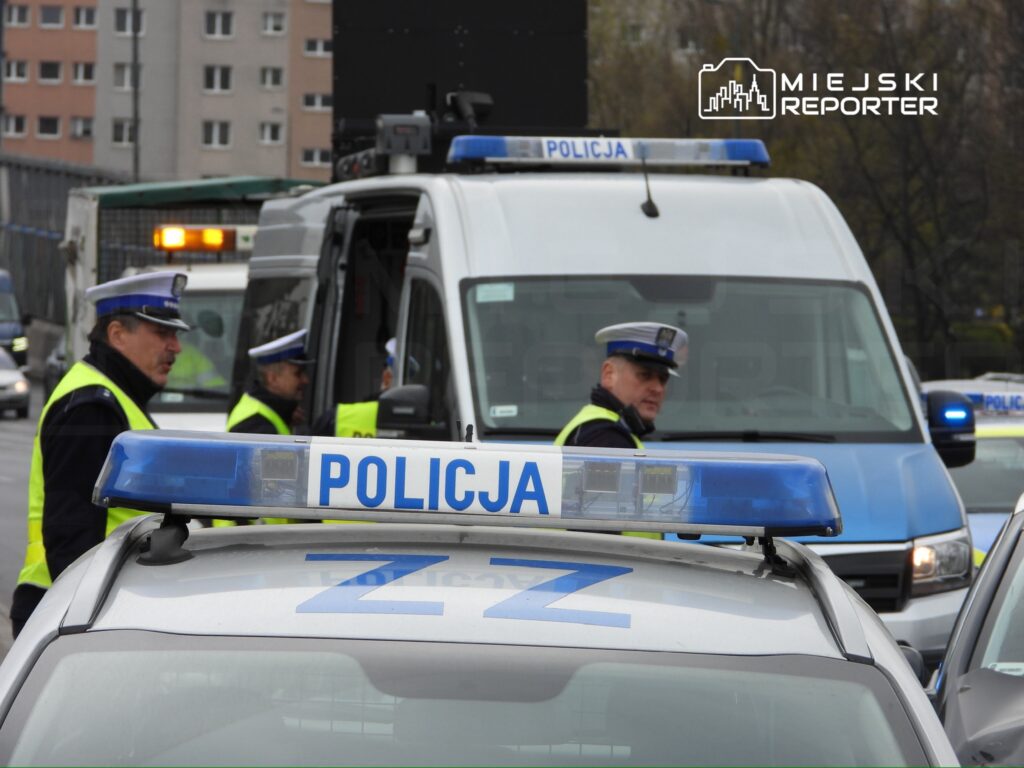 Grupa policjantów w kamizelkach odblaskowych rozmawia obok radiowozu na ruchliwej ulicy.