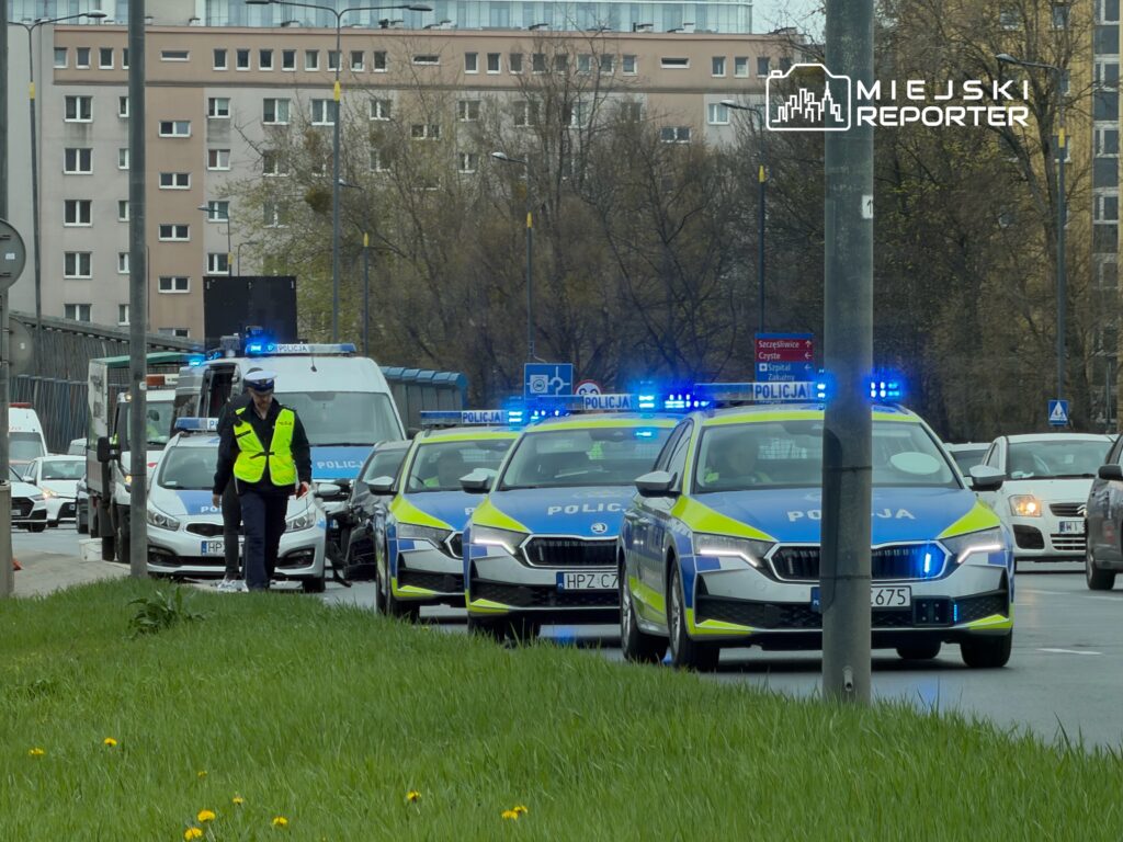 Policjant w kamizelce odblaskowej kieruje ruchem na ruchliwej ulicy, obok stoją policyjne samochody z włączonymi sygnałami.