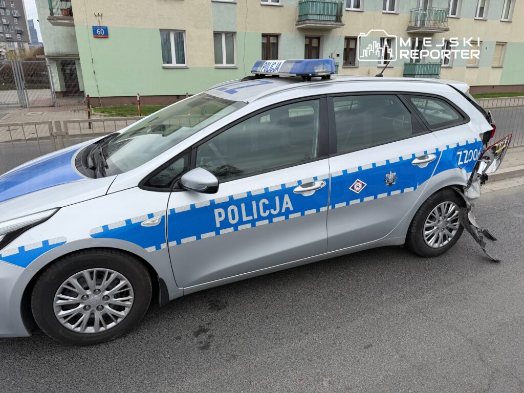 Policyjny samochód z uszkodzoną tylną częścią zaparkowany przy budynku mieszkalnym.