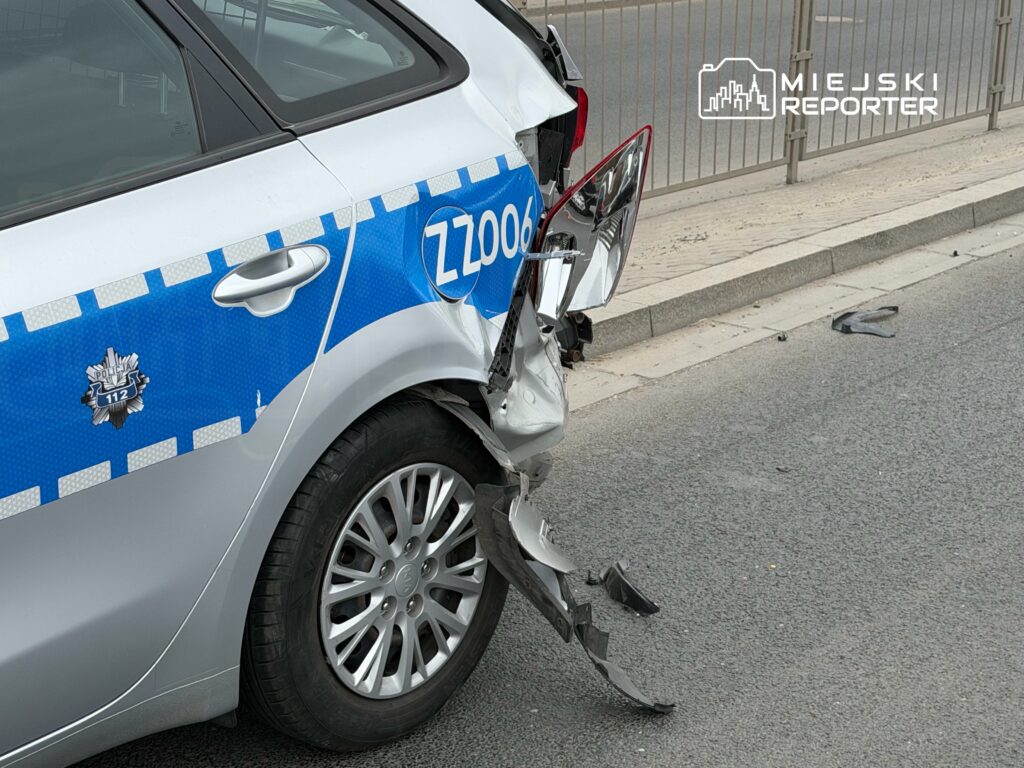 Uszkodzony radiowóz policyjny z wgniecioną tylną częścią na drodze, obok leżące fragmenty plastików.