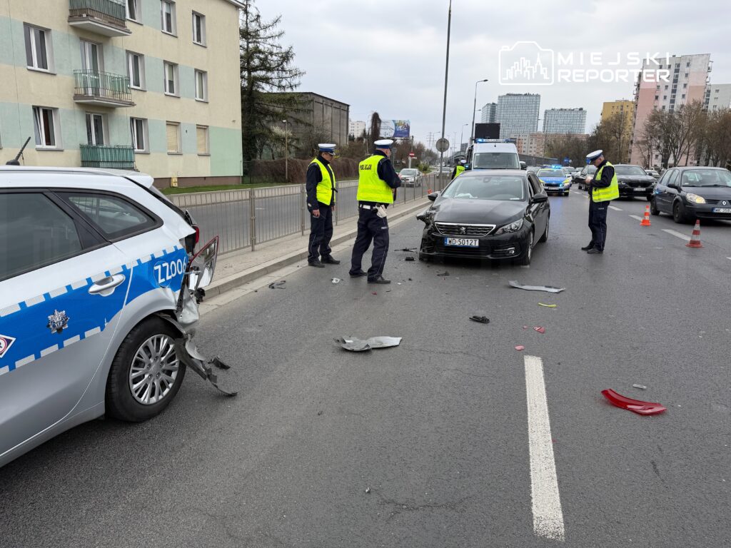 Policjanci w kamizelkach odblaskowych badają miejsce wypadku drogowego, w tle uszkodzony radiowóz i czarny samochód.
