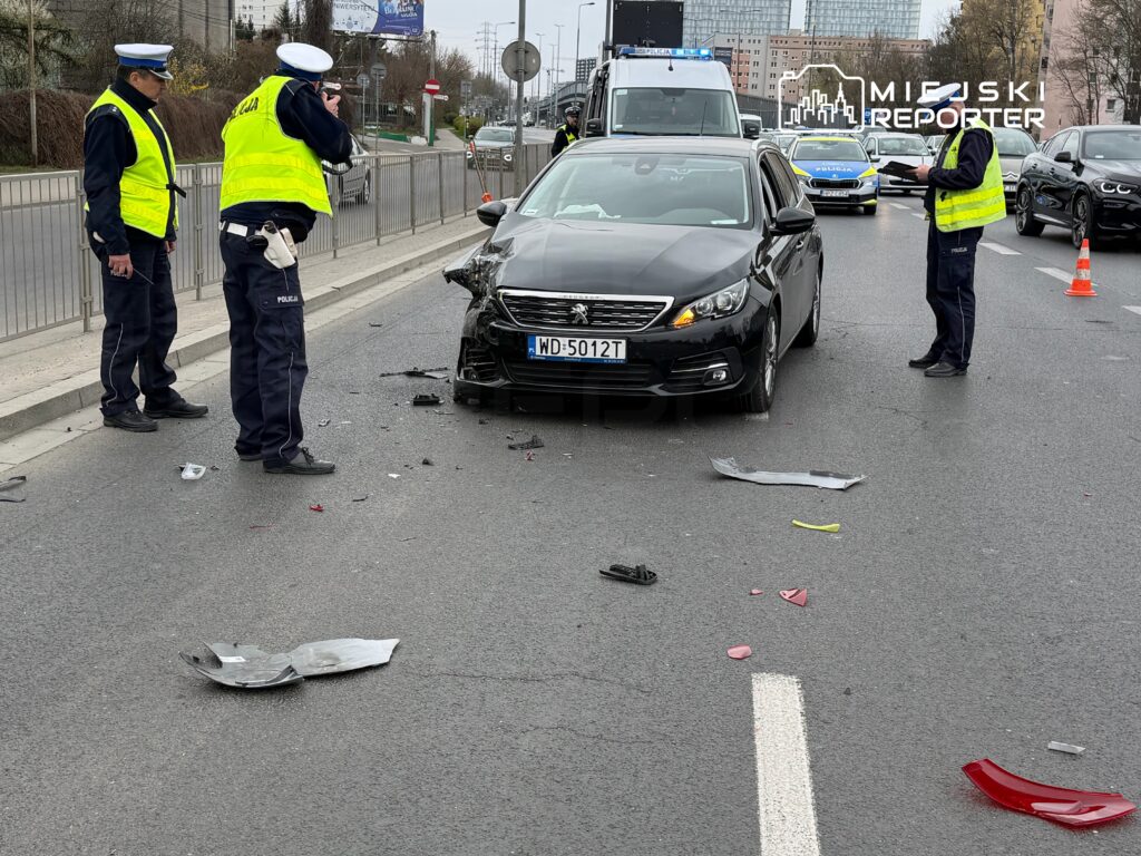 Policjanci w odblaskowych kamizelkach prowadzą czynności po kolizji drogowej z udziałem czarnego samochodu osobowego.