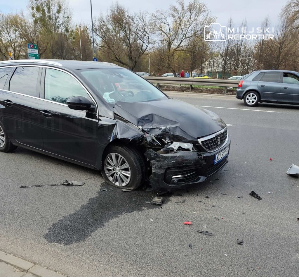Czarny samochód osobowy z uszkodzoną przednią częścią stoi na jezdni, obok leżą fragmenty wraku.