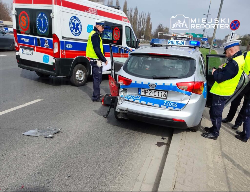Policyjny radiowóz z uszkodzeniem tyłu stoi na drodze, obok karetka pogotowia, a funkcjonariusze rozmawiają.