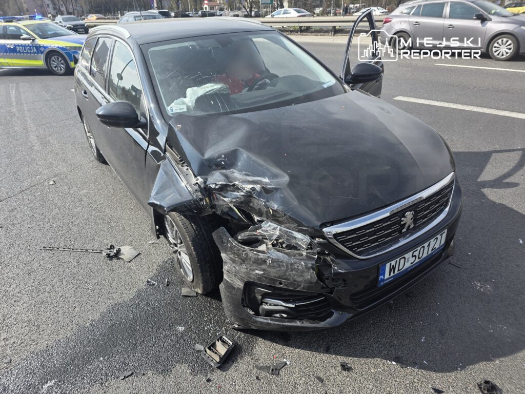 Czarny samochód osobowy z uszkodzonym przodem stoi na drodze, obok widać policyjny radiowóz.