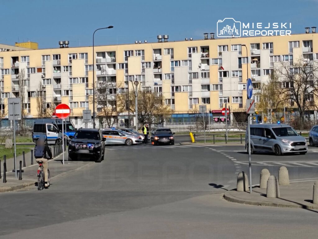 Mężczyzna na rowerze przejeżdża obok policyjnych samochodów blokujących skrzyżowanie w miejskim otoczeniu.
