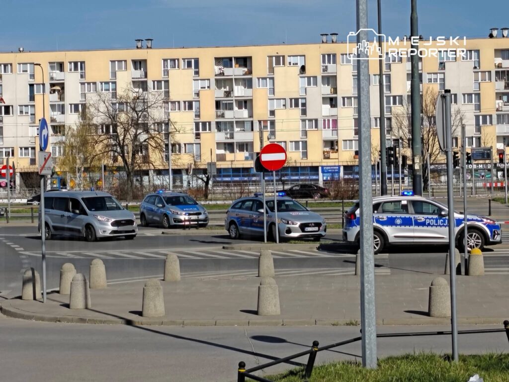 Samochód osobowy skręca w uliczkę, obok stoją dwa radiowozy policyjne w pobliżu bloków mieszkalnych.