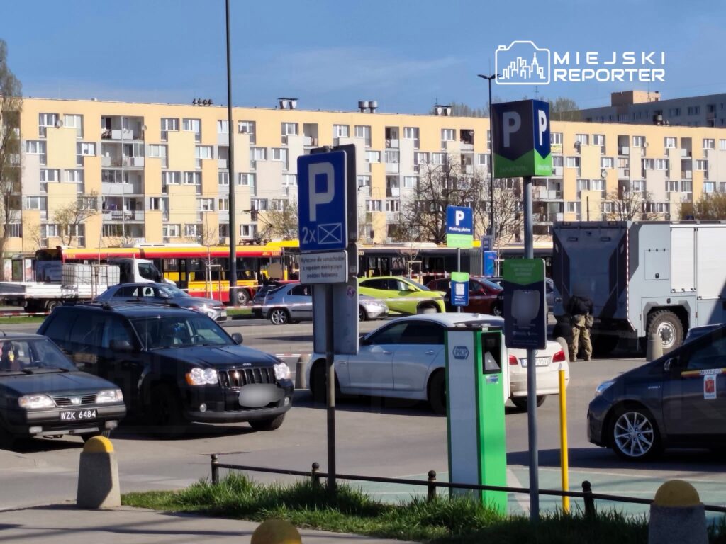 Ruchliwy parking z zaparkowanymi samochodami, w tle widać autobus oraz budynki mieszkalne.
