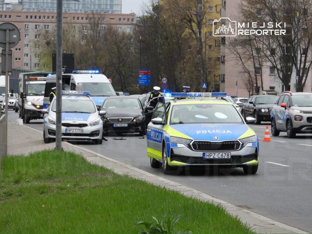 Policyjny radiowóz z włączonymi sygnałami świetlnymi zatrzymany na ruchliwej ulicy, obok inne pojazdy w korku.
